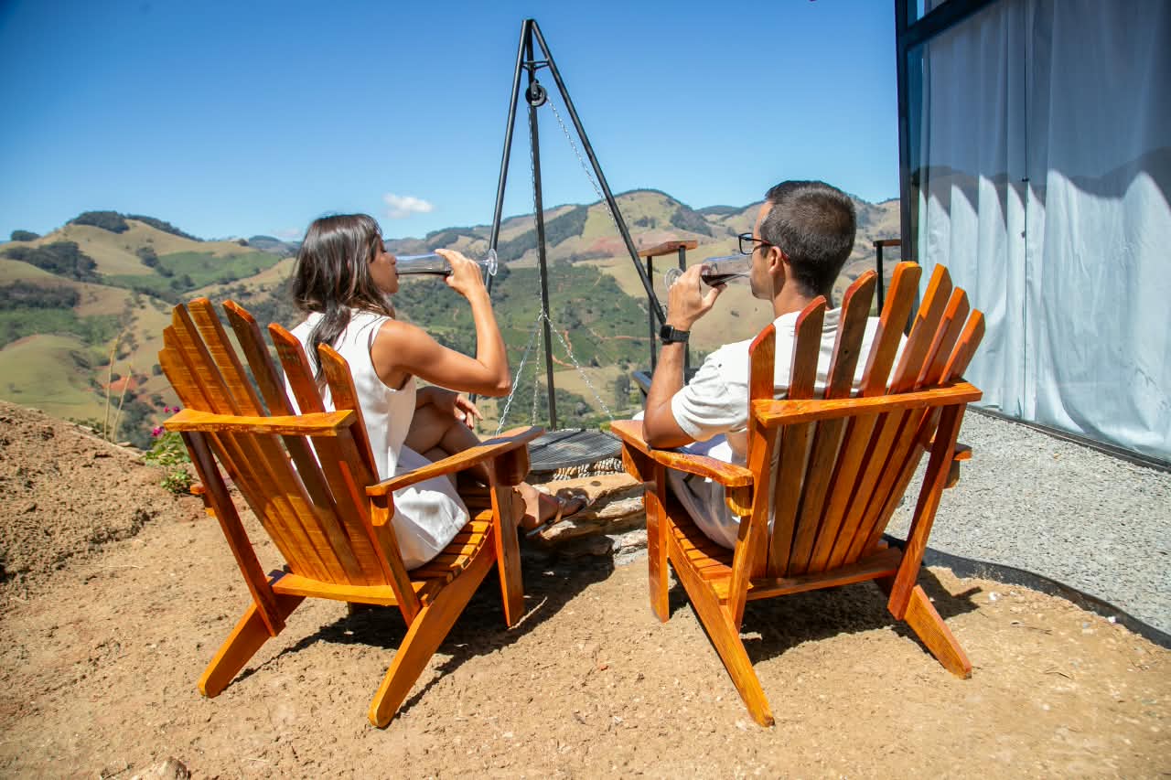 Casal brindando com vinho em cadeiras de madeira diante do vale de Cristina
