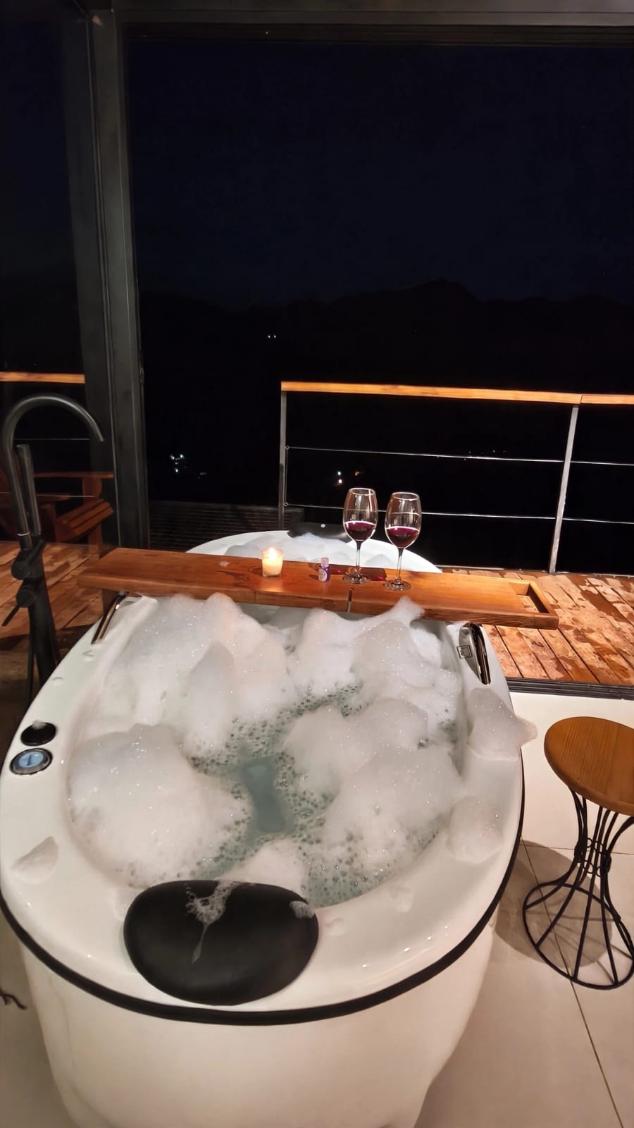 Banheira de hidromassagem com espuma, duas taças de vinho e vela na varanda à noite